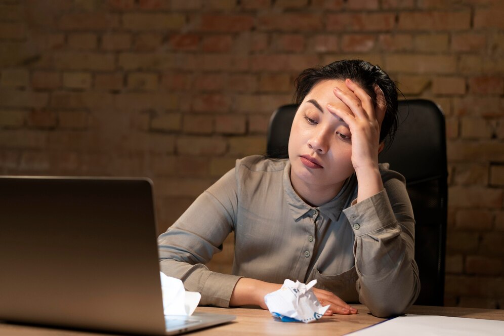 woman sits at a desk, resting her head in her hands, conveying a sense of stress or deep contemplation