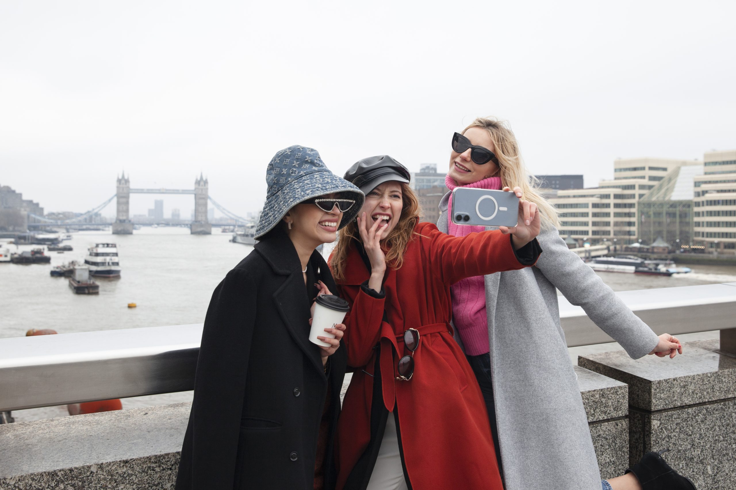 Three friends pose for a joyful selfie on a London bridge, with Tower Bridge in the background